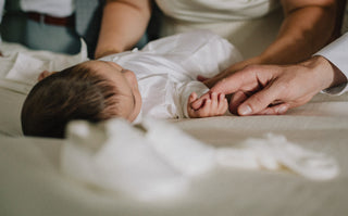 baby lays down on bedsheet while holding an adult's finger