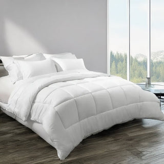 White comforter and pillows on a bed in a room with large windows showing greenery outside.