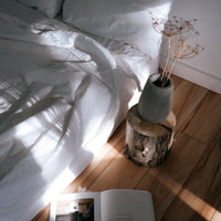 Vase with dried plants on a wooden stool next to a bed with white bedding.